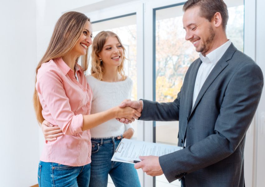 Two smiling tenants shake the hand of a property manager in a suit, who holds a clipboard.